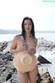 A woman in a straw hat standing on a rock by the ocean.