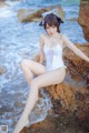 A woman in a white bathing suit sitting on a rock by the ocean.